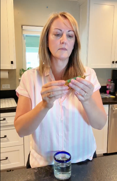 A woman in a kitchen looks intently at a small object she holds over a glass of water.