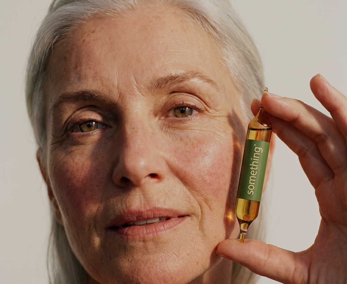 A close-up of a woman with grey hair holding a small glass ampoule with a green 'something' label.