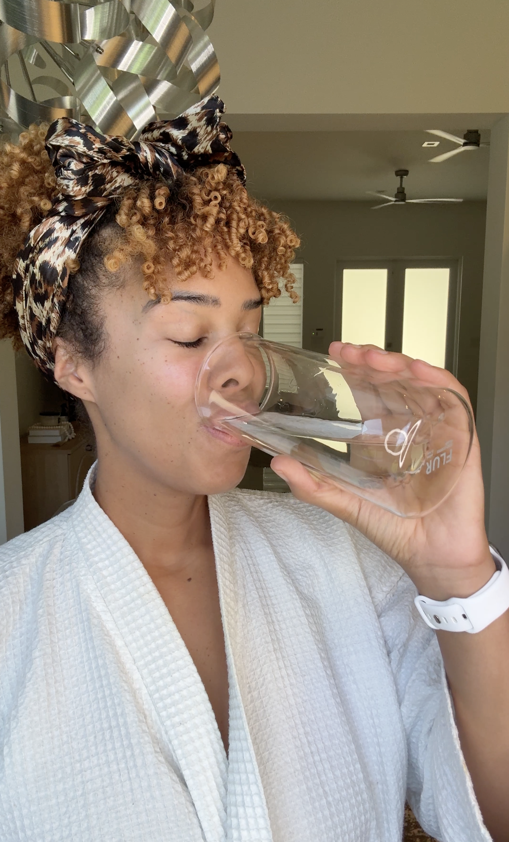A person wearing a white robe and leopard-print headscarf drinks a glass of water with their eyes closed.