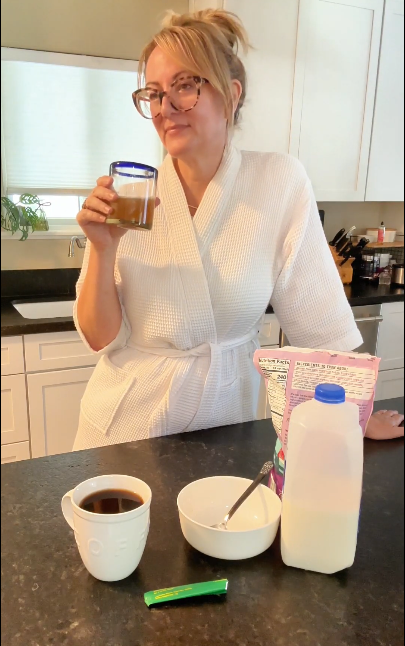 A woman in a white robe stands at a kitchen counter with breakfast items.