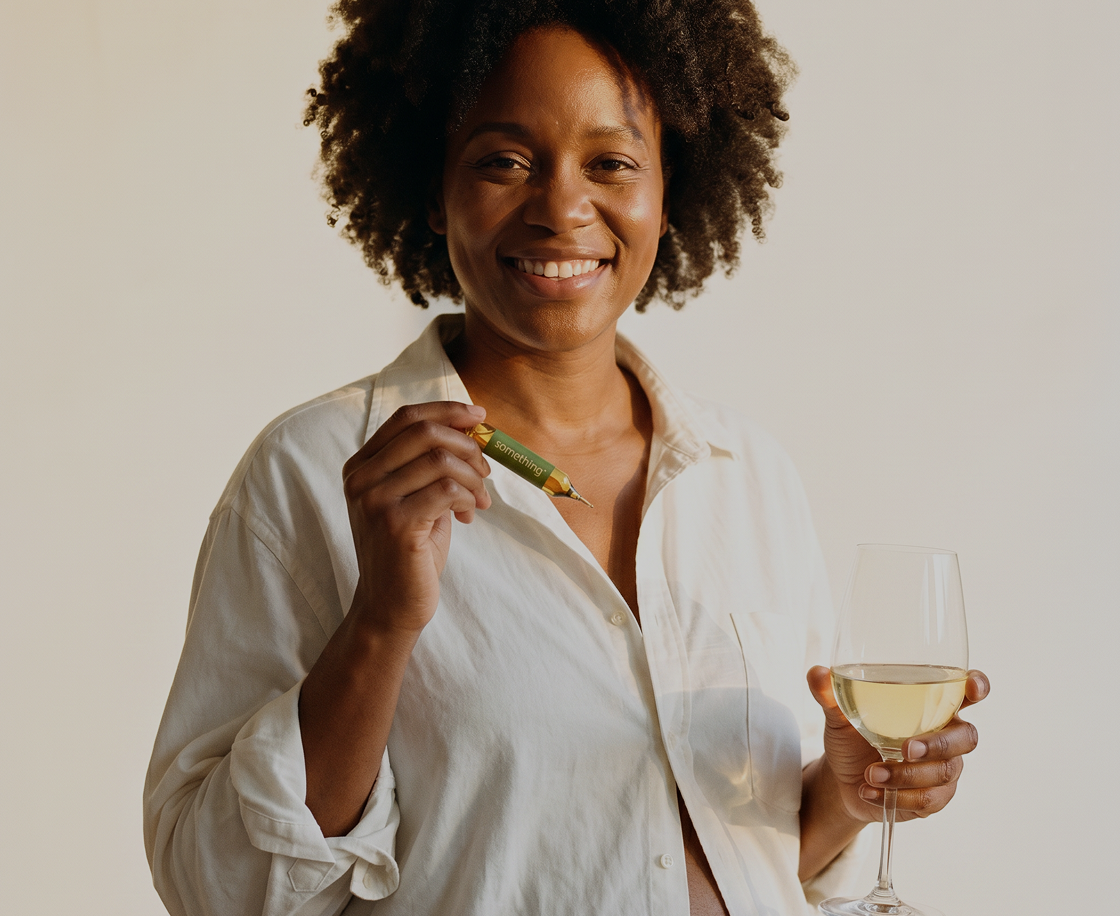 A smiling woman in a white shirt holds a small green product and a glass of white wine.