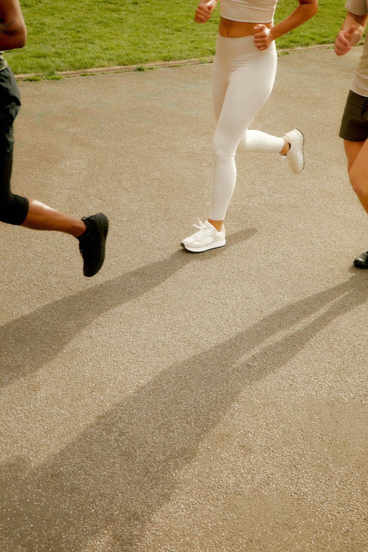 The lower bodies of three people in athletic wear running on a paved path next to grass.