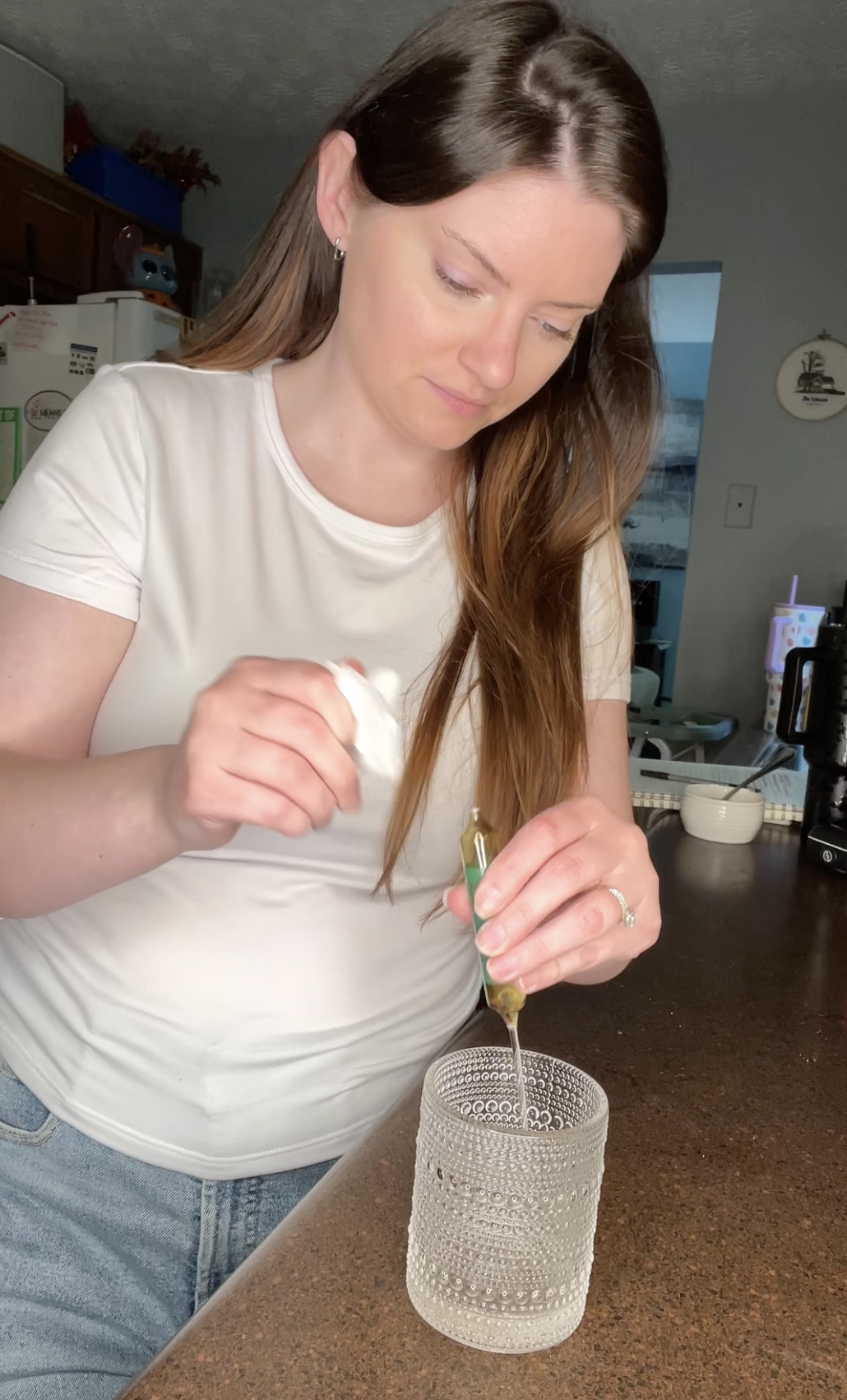 A woman in a white t-shirt and jeans squeezes liquid from a dropper into a textured glass.