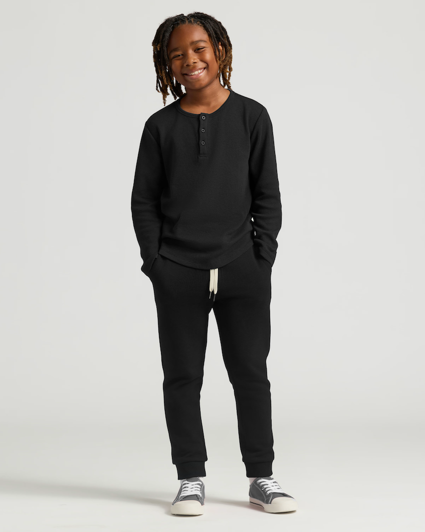 Smiling child in black outfit with hands in pockets, standing against a plain background.