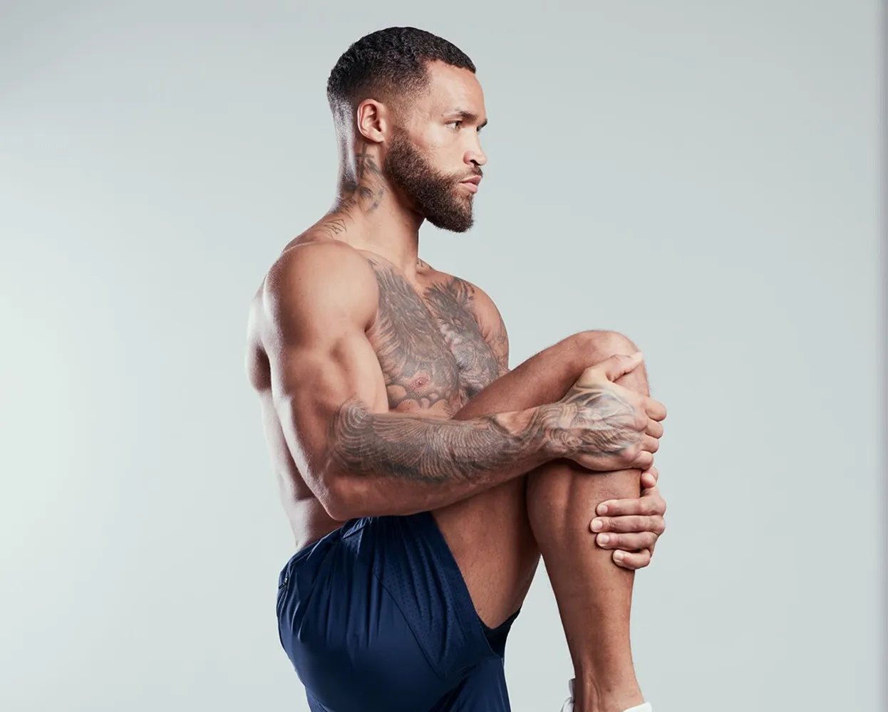 Man with tattoos stretching, holding one knee up, wearing blue shorts.