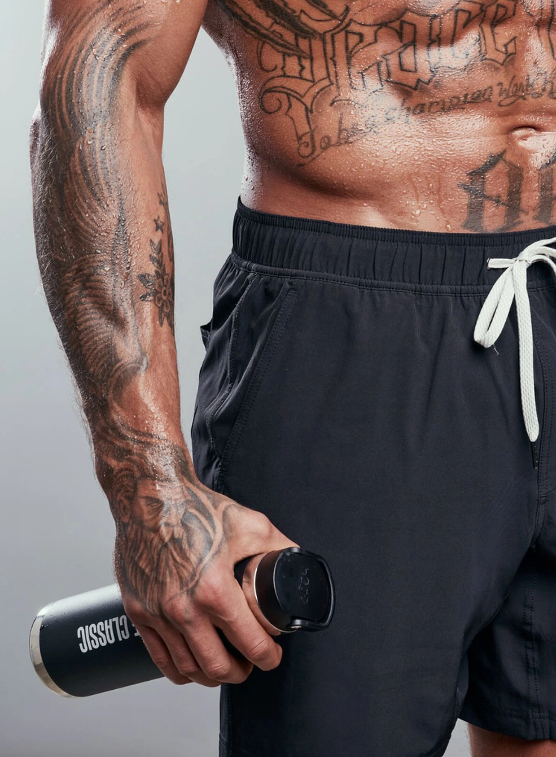 Tattooed person holding a black water bottle wearing black shorts.