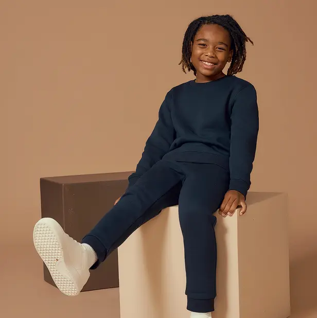 Child in navy tracksuit sitting on a beige block, smiling.