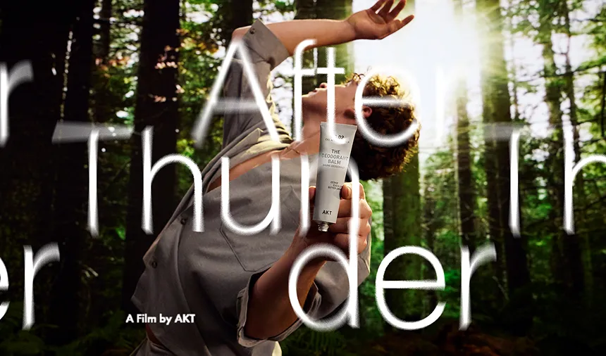 A person poses in a sunlit forest, holding a tube of 'The Deodorant Balm' by AKT.