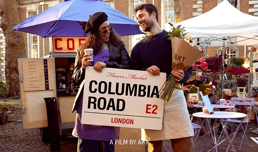 A smiling man and woman hold a 'Columbia Road' sign and a bouquet at an outdoor market.