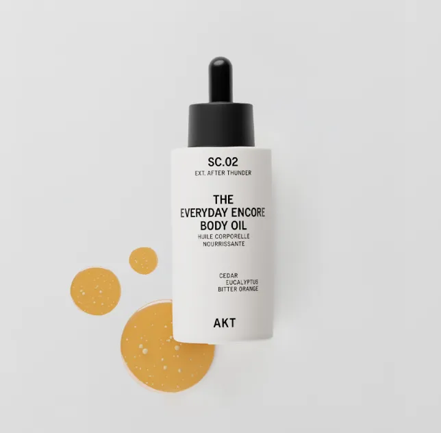 A white bottle of AKT body oil with a black dropper cap, beside three drops of golden oil.