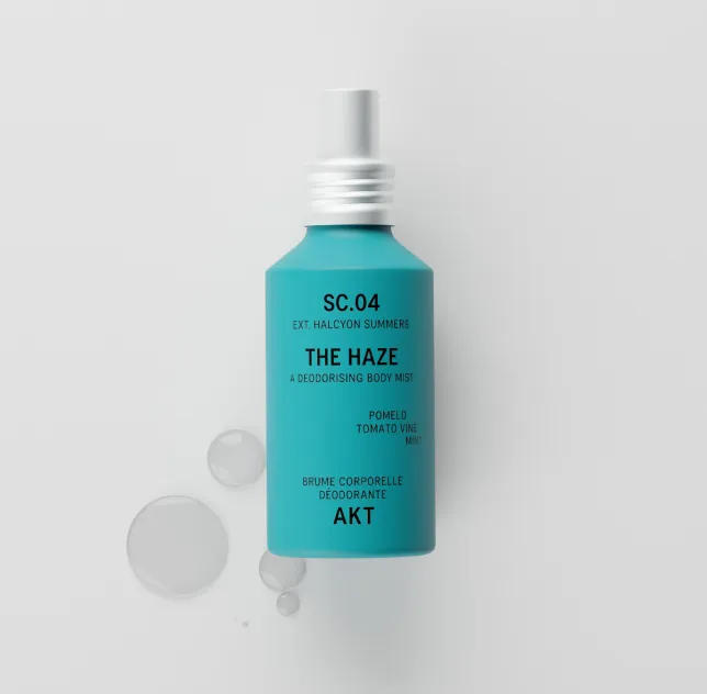 A teal bottle of AKT's The Haze deodorising body mist on a white surface with clear droplets.