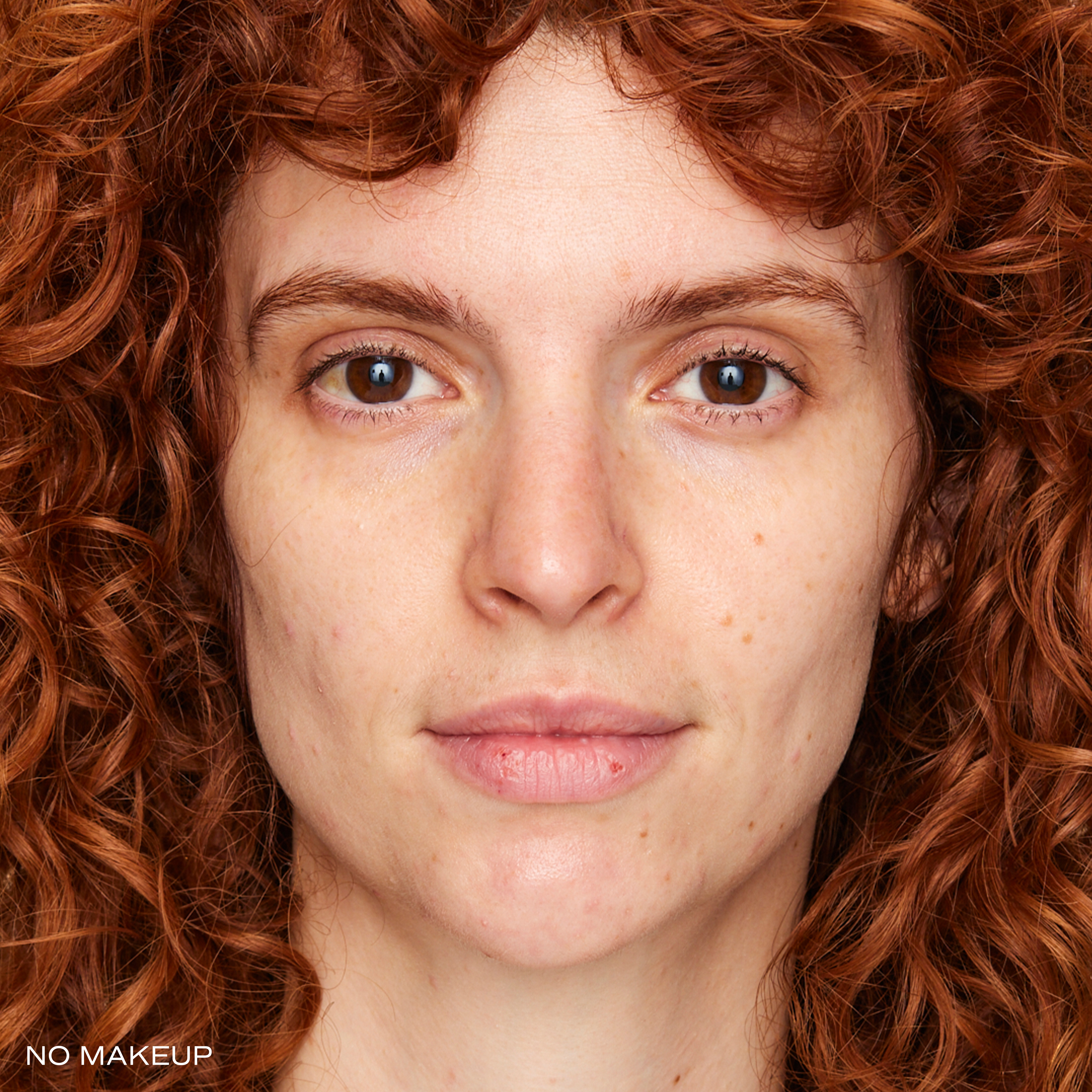 A close-up of a woman with curly red hair and brown eyes, not wearing any makeup.