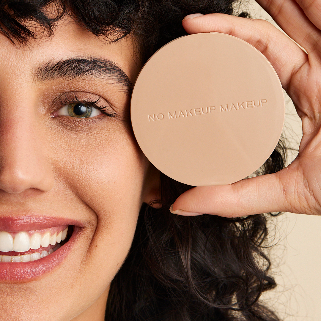 A smiling woman with dark curly hair holds a beige makeup compact, with 'NO MAKEUP MAKEUP' on it, over her eye.