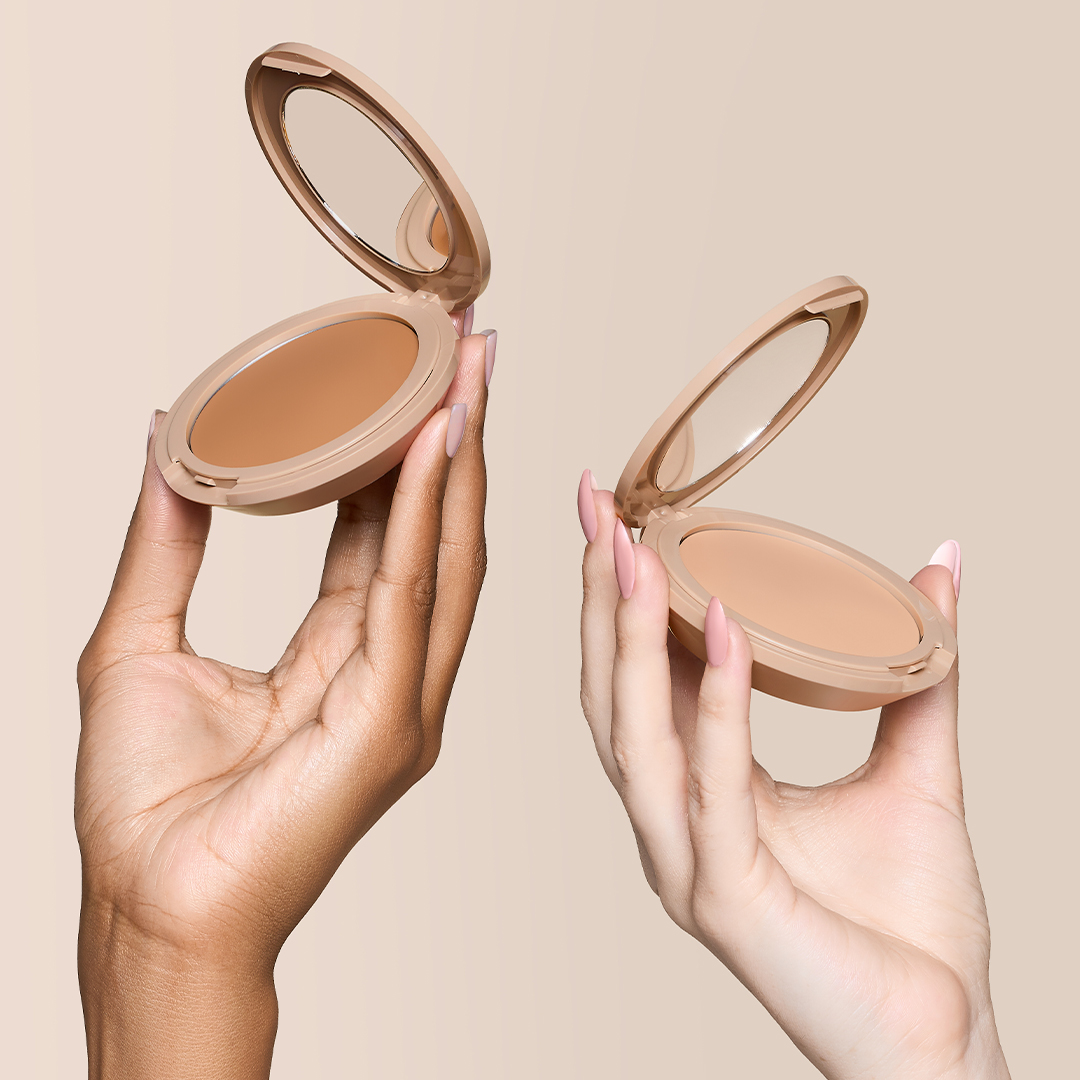 Two hands with different skin tones each hold an open makeup compact with foundation inside.