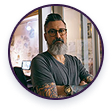 A man with a grey beard, glasses, and tattooed arms, stands with his arms crossed in an office.