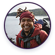 A smiling person with dreadlocks and a backpack, wearing a red jacket and headband outdoors.