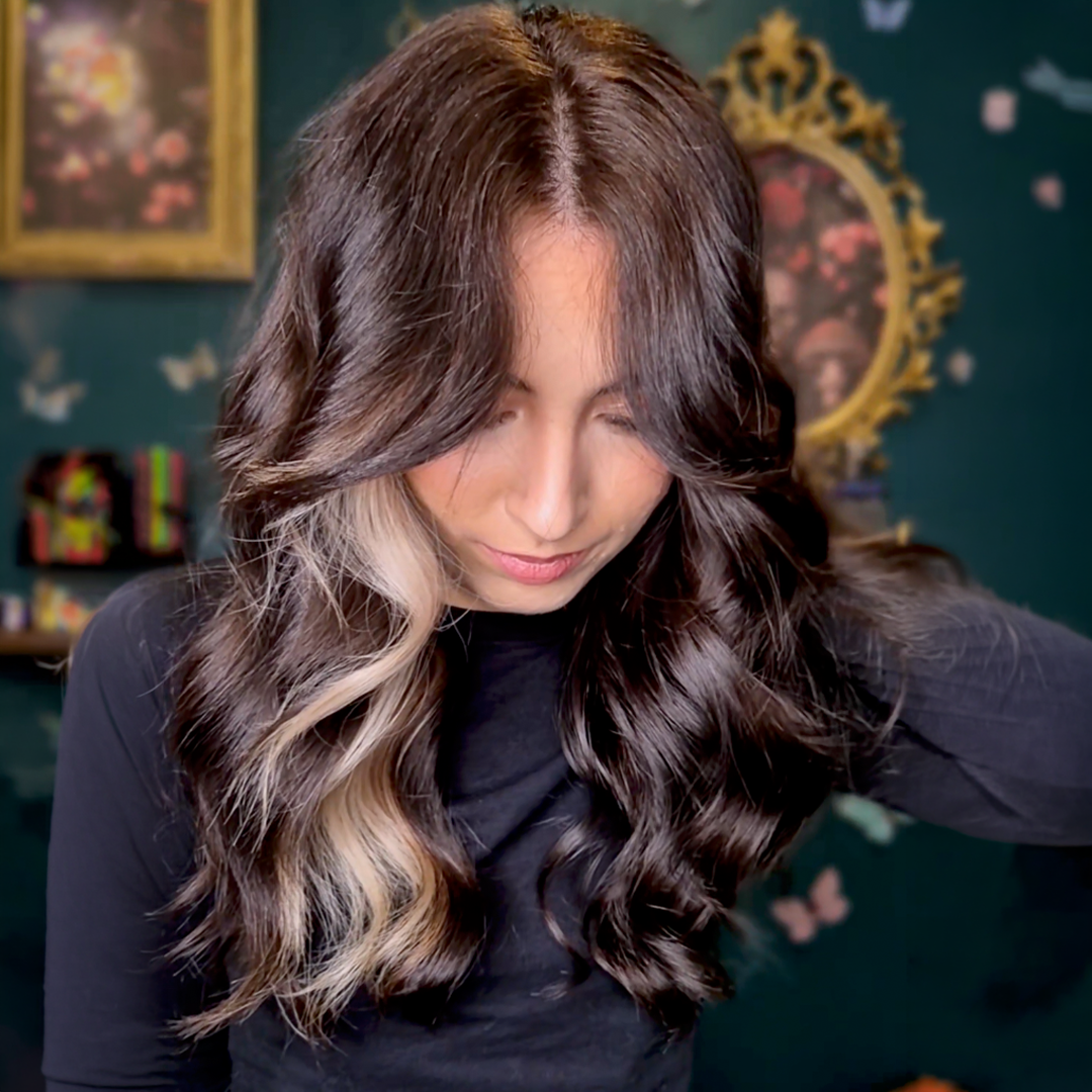A woman with long, wavy, dark brown hair with a prominent blonde streak in the front looks down.
