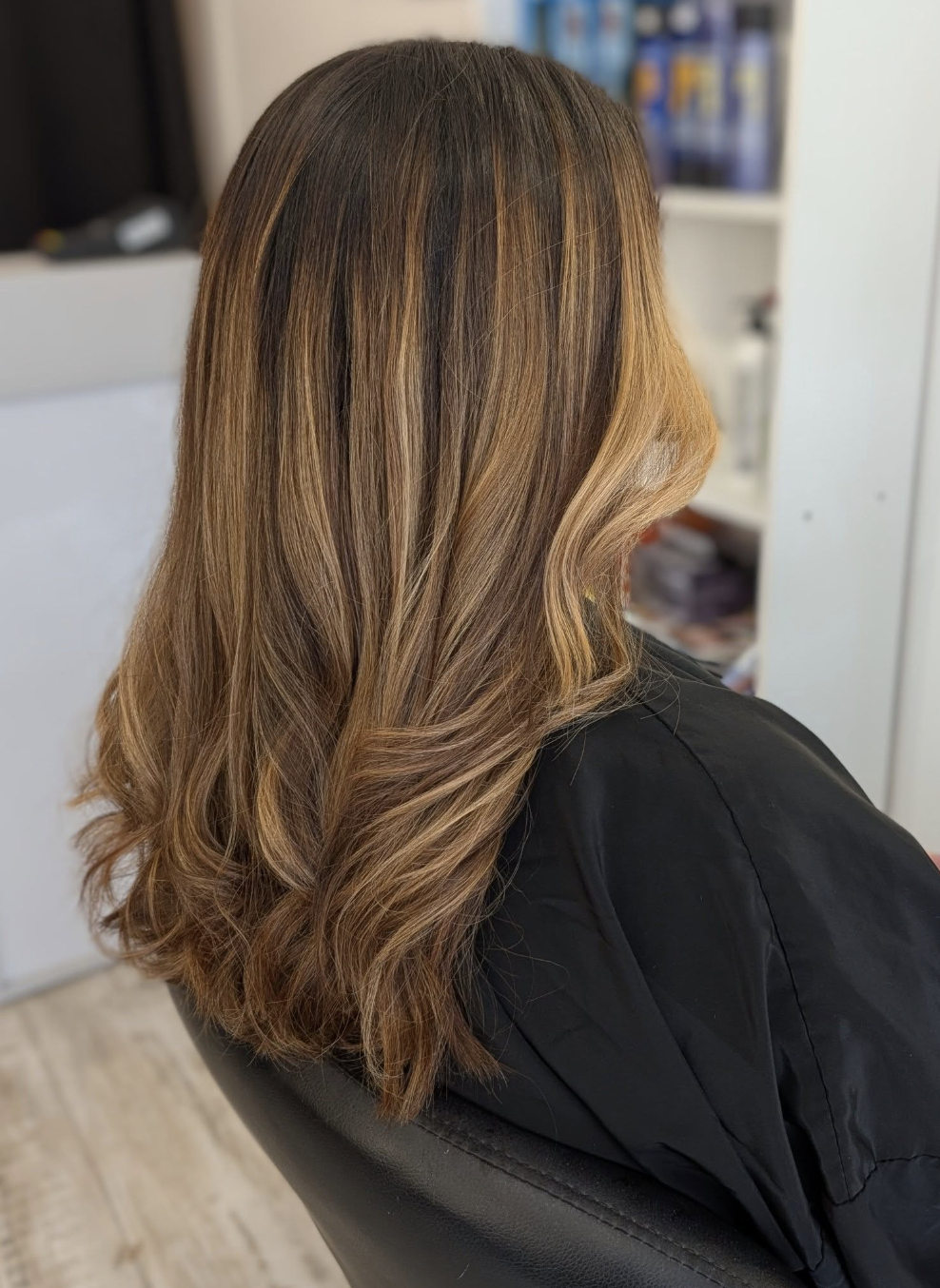 The back of a woman with long, wavy brown hair with blonde highlights wearing a salon cape.