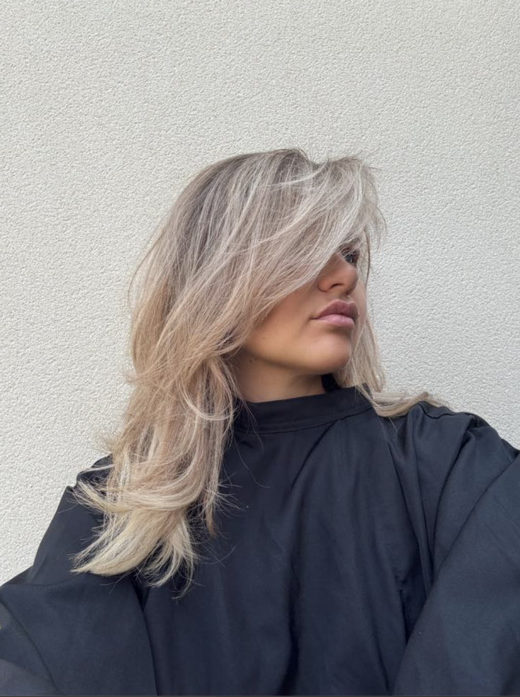 A woman with layered blonde hair covering her face, wearing a black salon cape against a white wall.