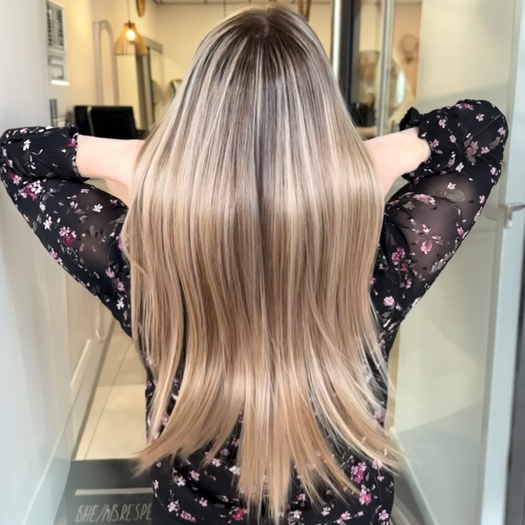 The back of a woman with long, straight, highlighted blonde hair, holding her hands behind her head.
