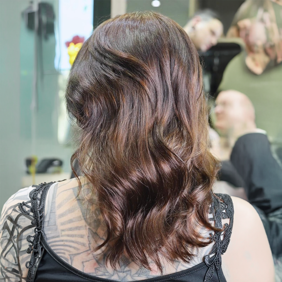 Rear view of a person with wavy, shoulder-length brown hair and tattoos on their back and shoulders.