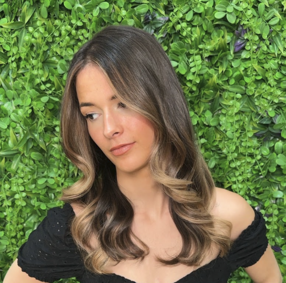 A woman with long, wavy, highlighted hair poses in front of a wall of green foliage.