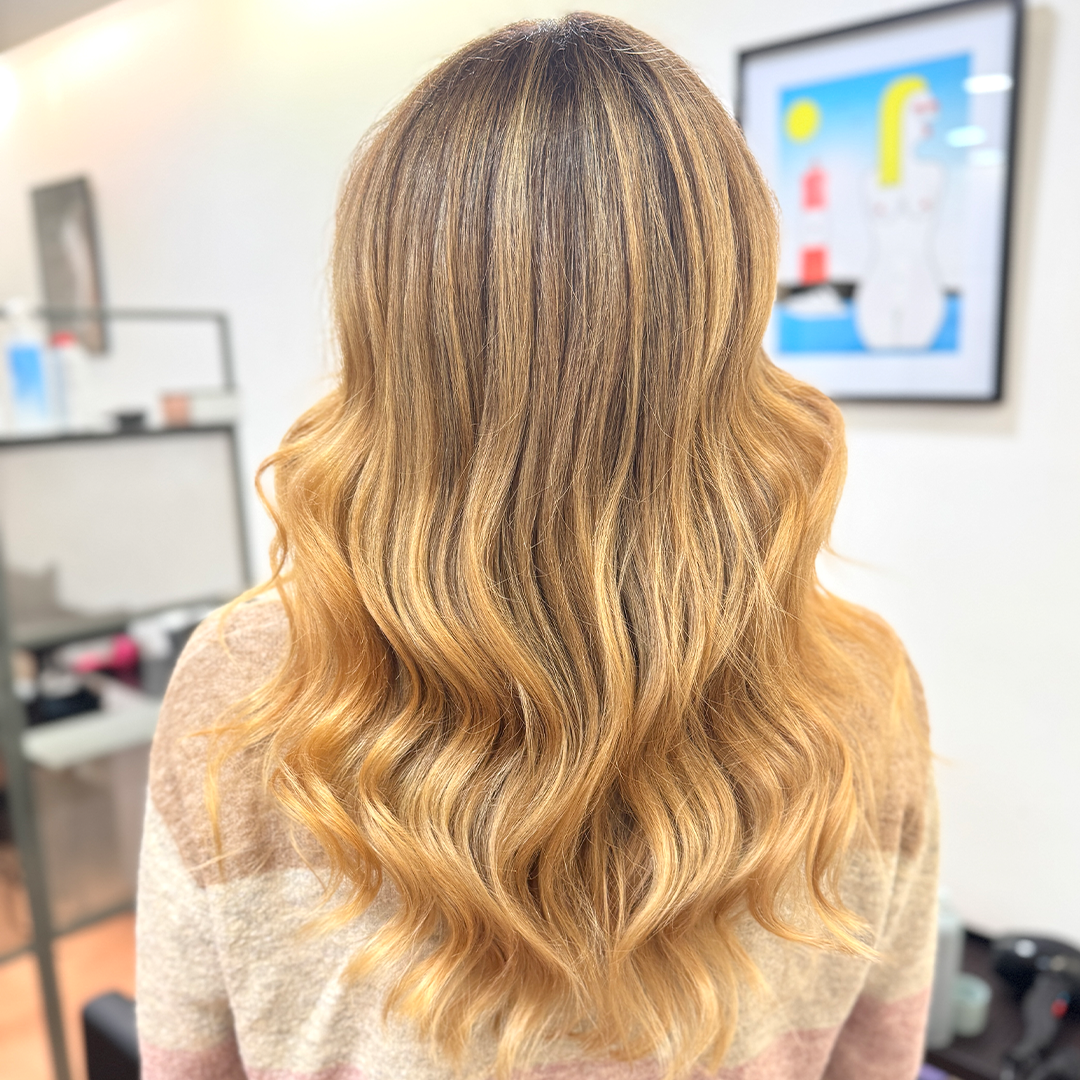 The back of a person with long, wavy, honey-blonde hair with highlights, wearing a striped sweater.