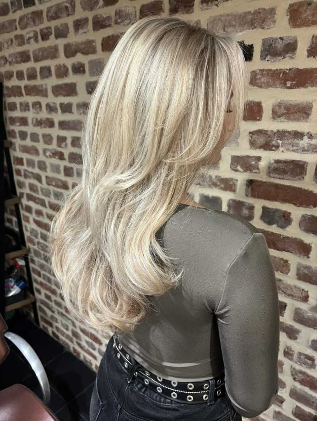 Back view of a person with long, layered blonde hair standing in front of a brick wall.