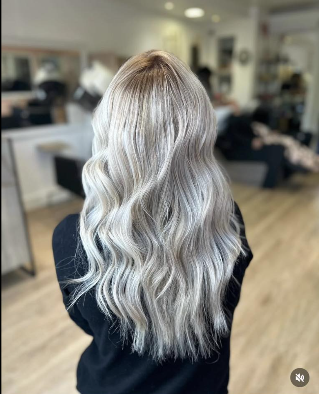 The back of a person with long, wavy, cool-toned blonde hair with dark roots in a salon.
