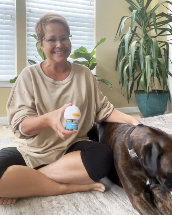 Smiling person sitting with a dog, holding a small container, indoors with plants.
