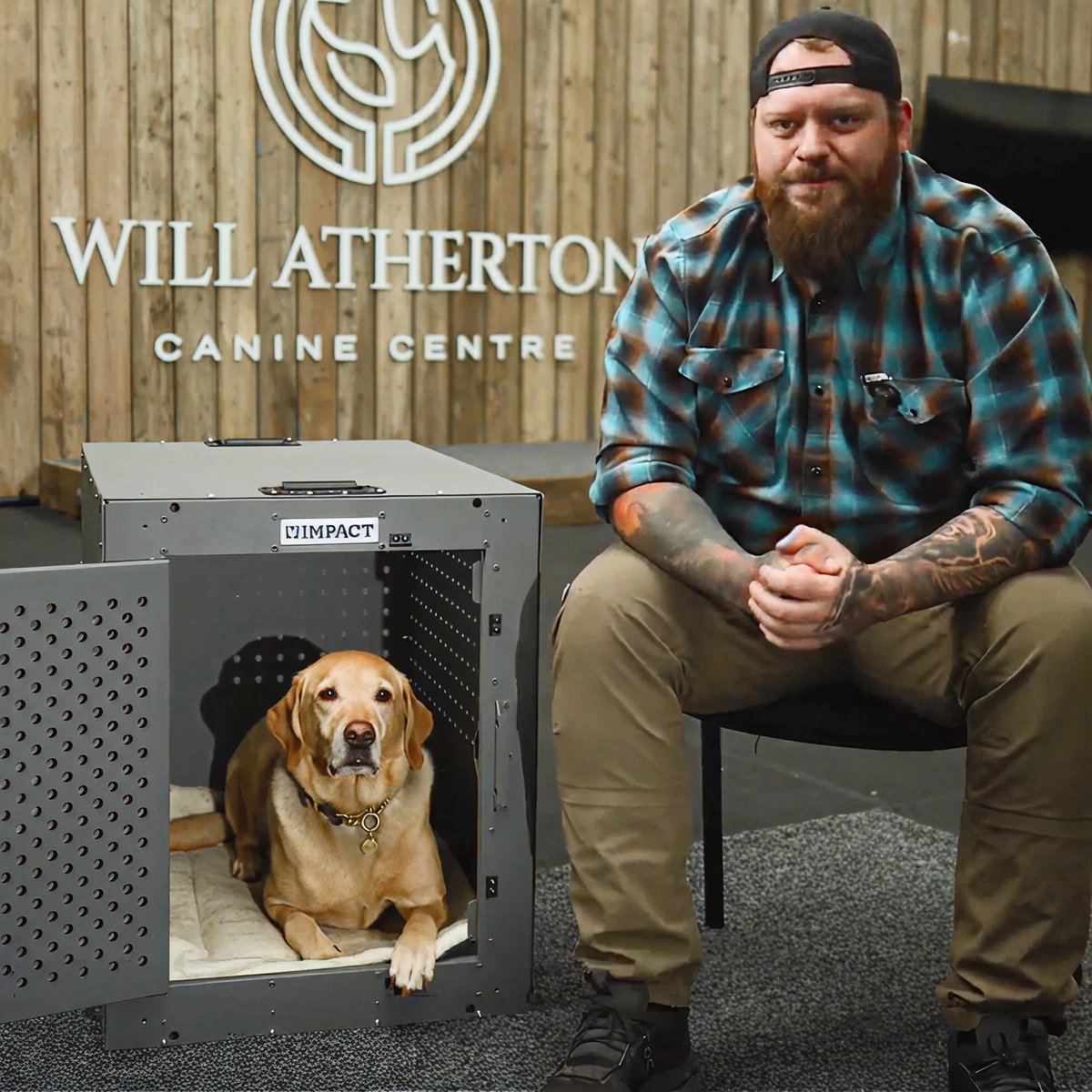 A man with a beard sits next to a yellow Labrador retriever lying in an open crate.