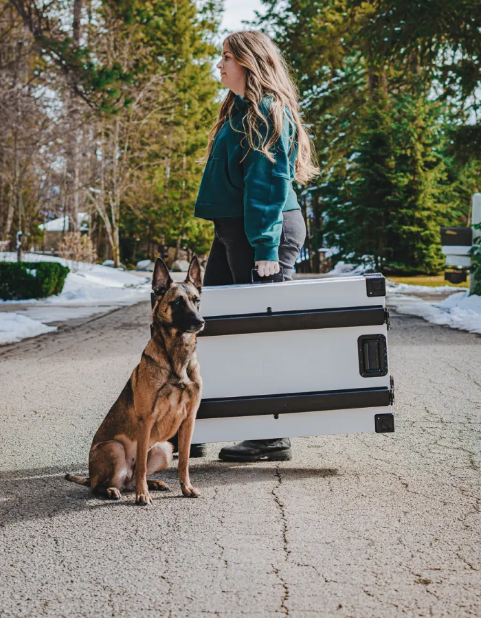 A woman stands on a paved road with a Belgian Malinois dog, holding a large folded object.