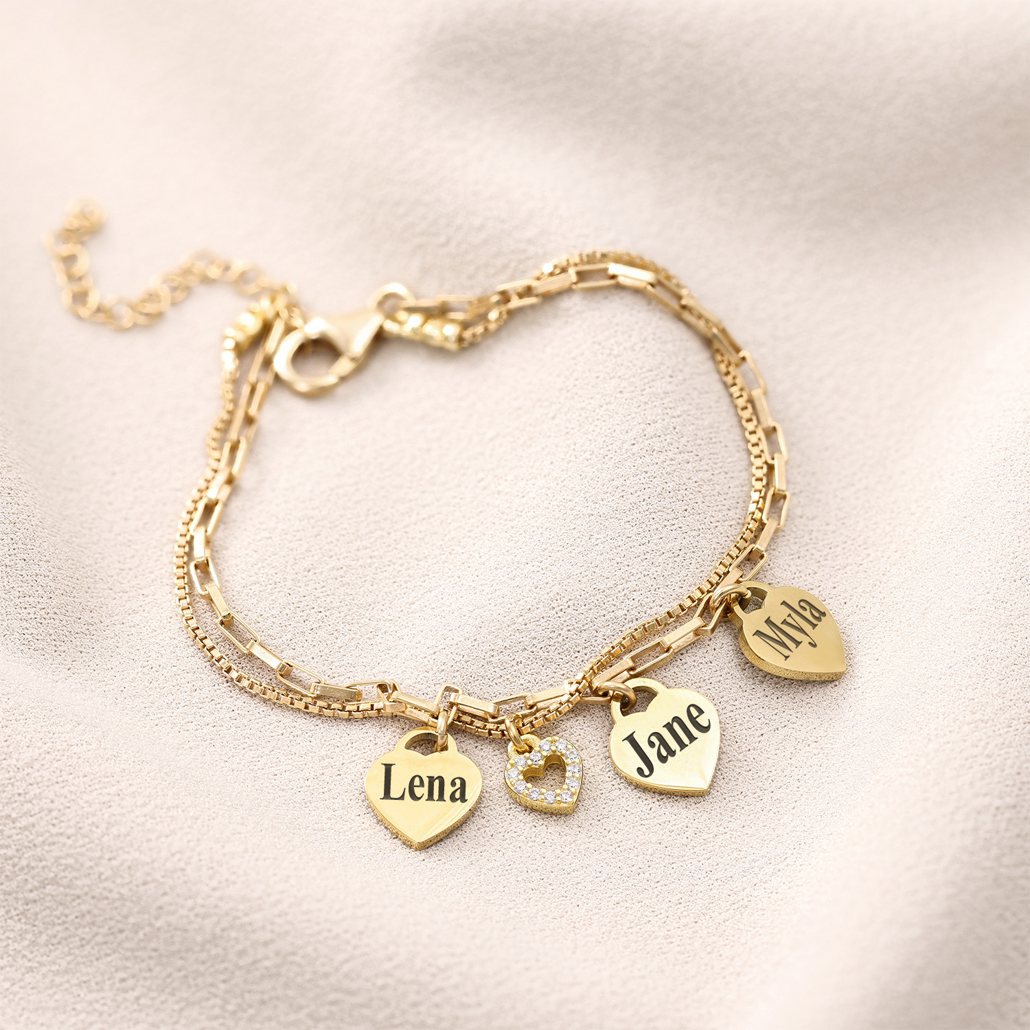 Gold bracelet with heart-shaped charms engraved with names on a soft fabric background.