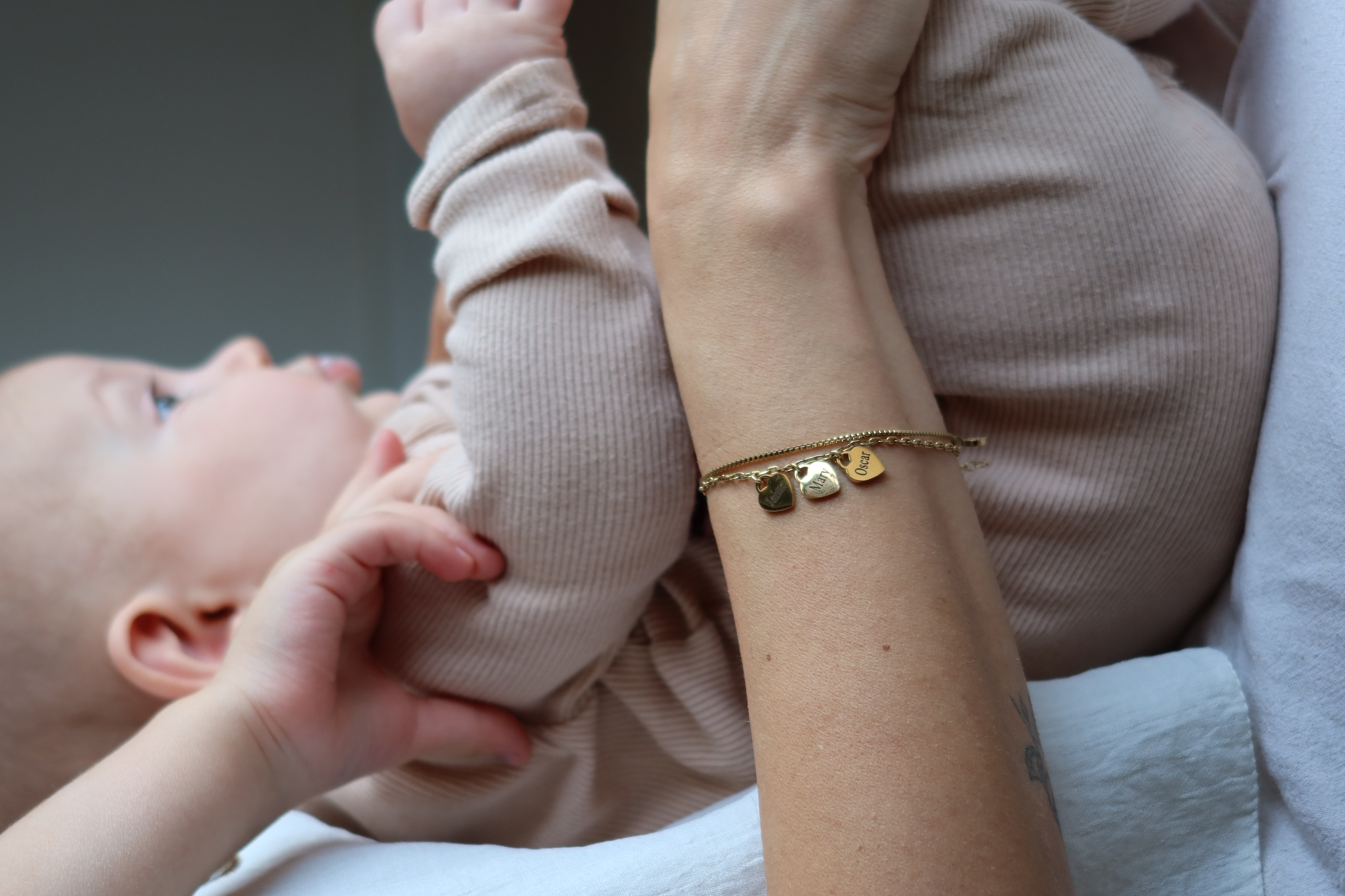 Baby in beige outfit held by an adult wearing a bracelet with name charms.