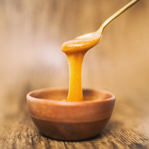 A close-up of thick golden honey dripping from a spoon into a small wooden bowl on a wooden surface.