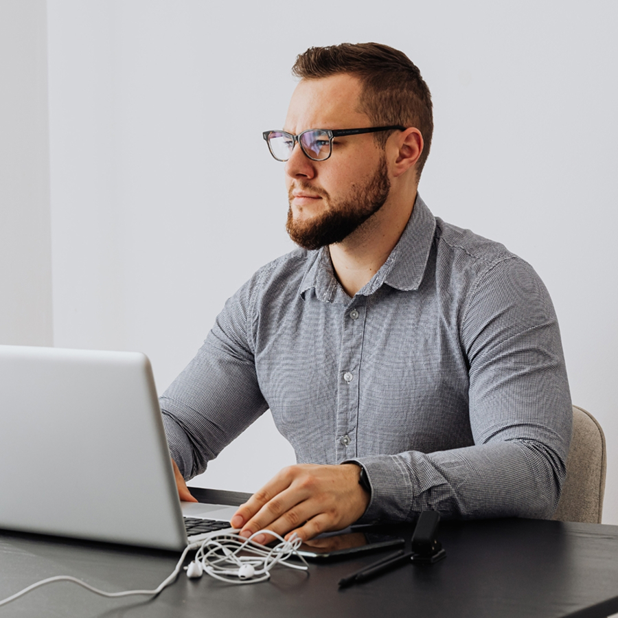 A bearded man with glasses in a grey button-down shirt sits at a desk using a laptop.