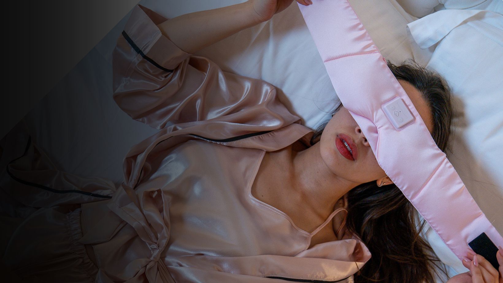 A woman in pink satin pajamas lies in bed putting on a pink electronic sleep mask.