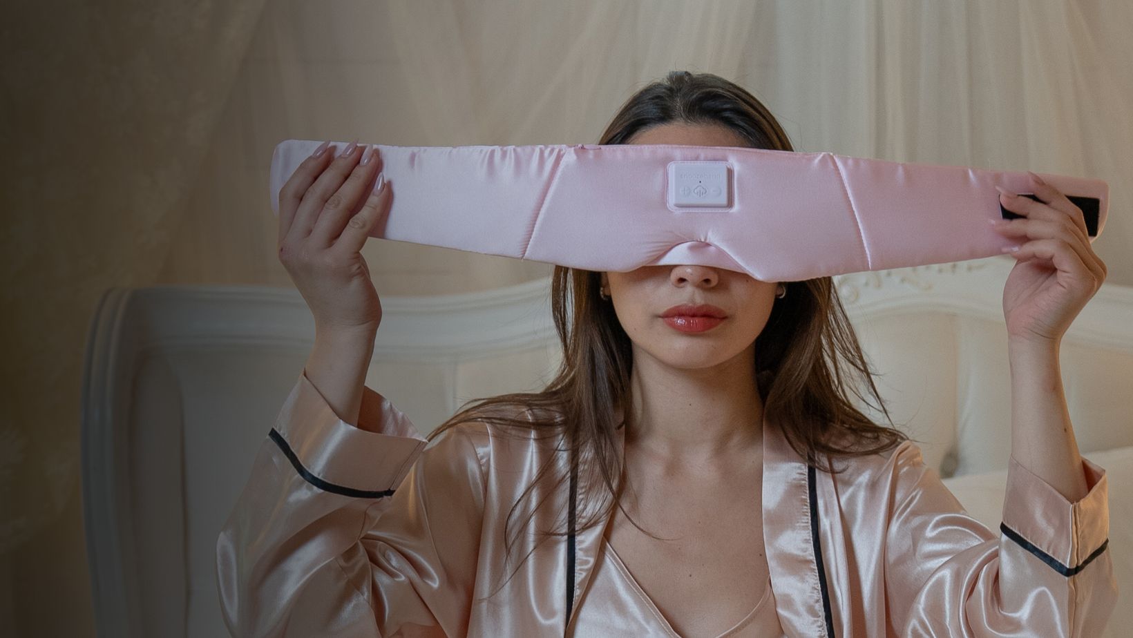 A woman in a pink satin robe holds a large, pink electronic eye massager over her eyes.