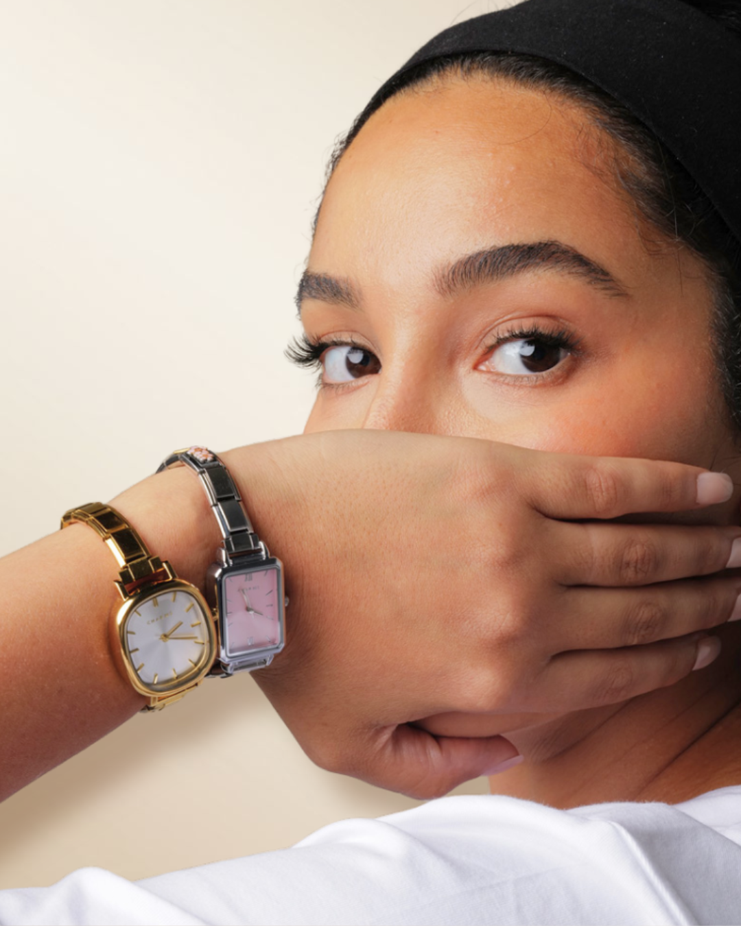 A woman with a black headband covers her mouth, revealing two watches on her wrist.