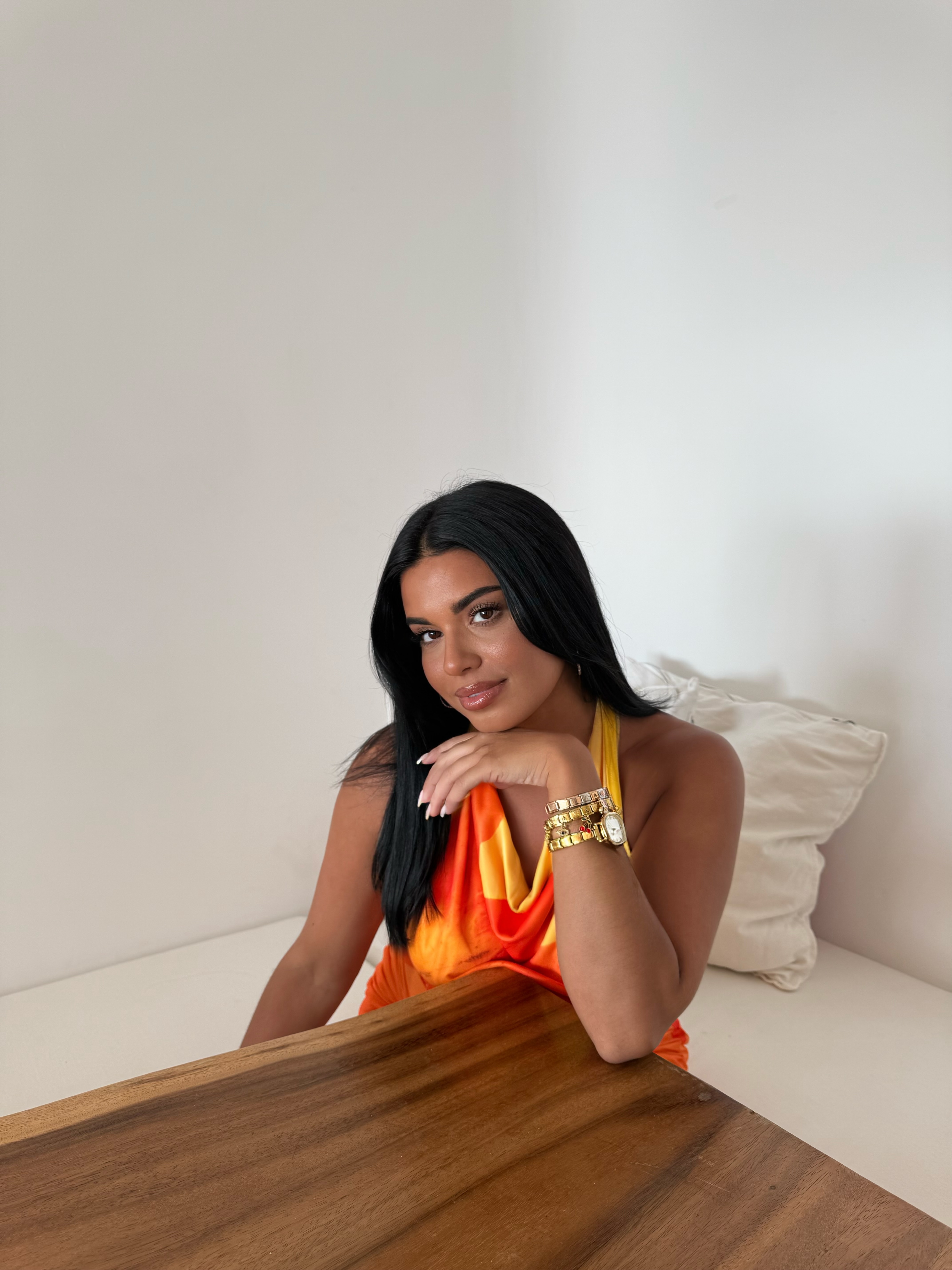 A woman with long dark hair in an orange and yellow top rests her chin on her hand at a table.