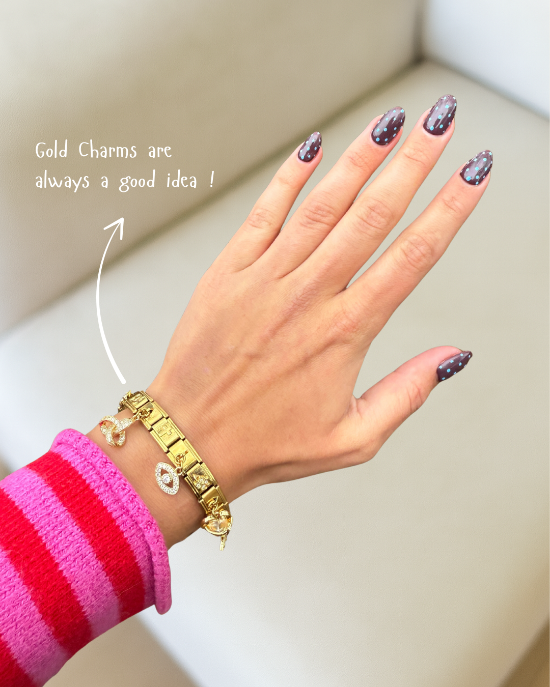 A woman's hand wearing a gold charm bracelet and showing off a dark manicure with blue polka dots.