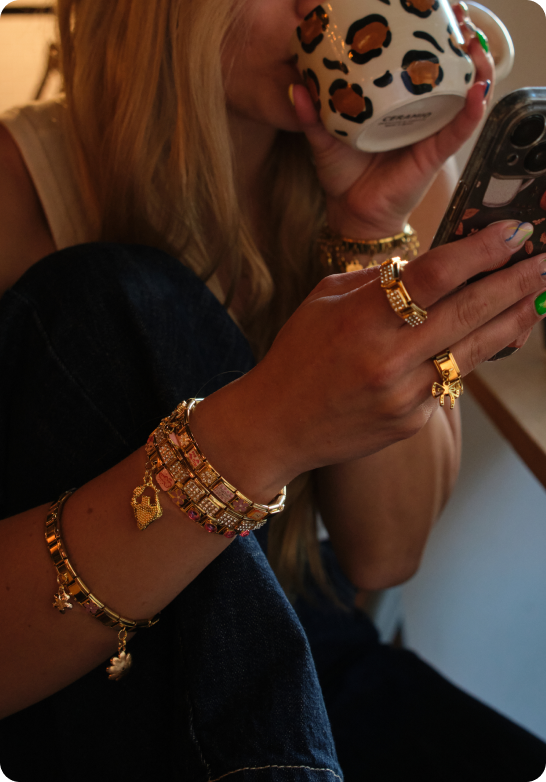 A person wearing multiple gold bracelets and rings drinks from a leopard-print mug while holding a phone.