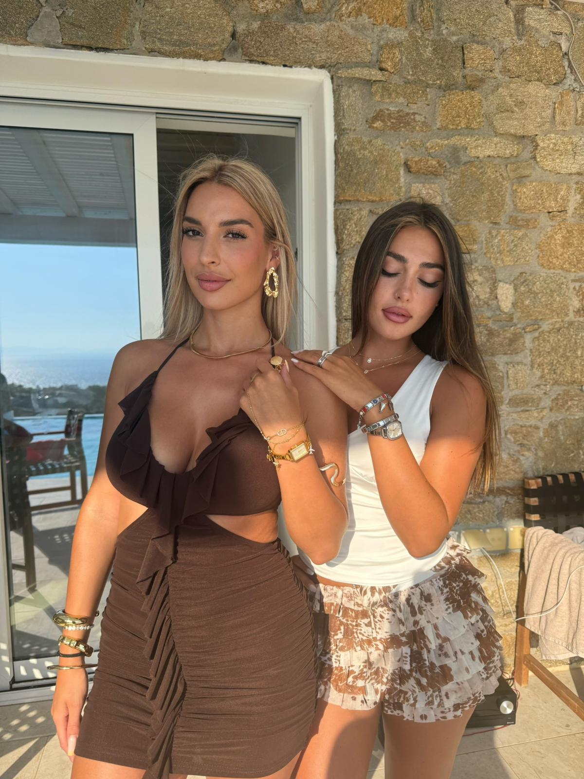 Two women pose together on a balcony with a stone wall and a view of the ocean.