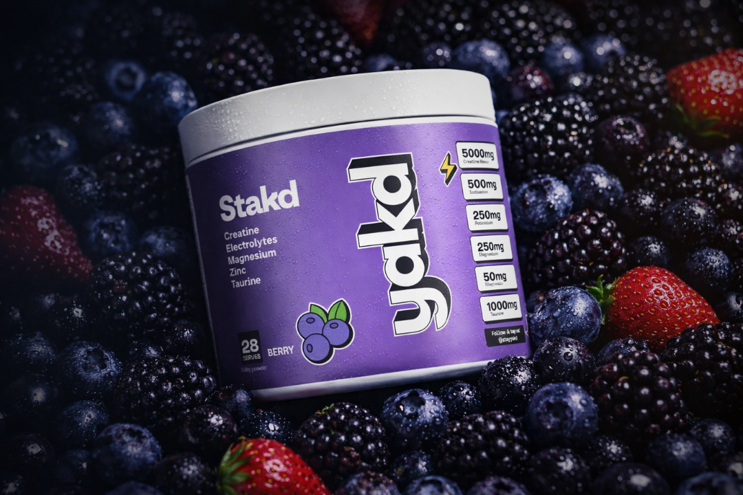 A purple container of Stakd Jakd supplement powder sits on a bed of fresh mixed berries.