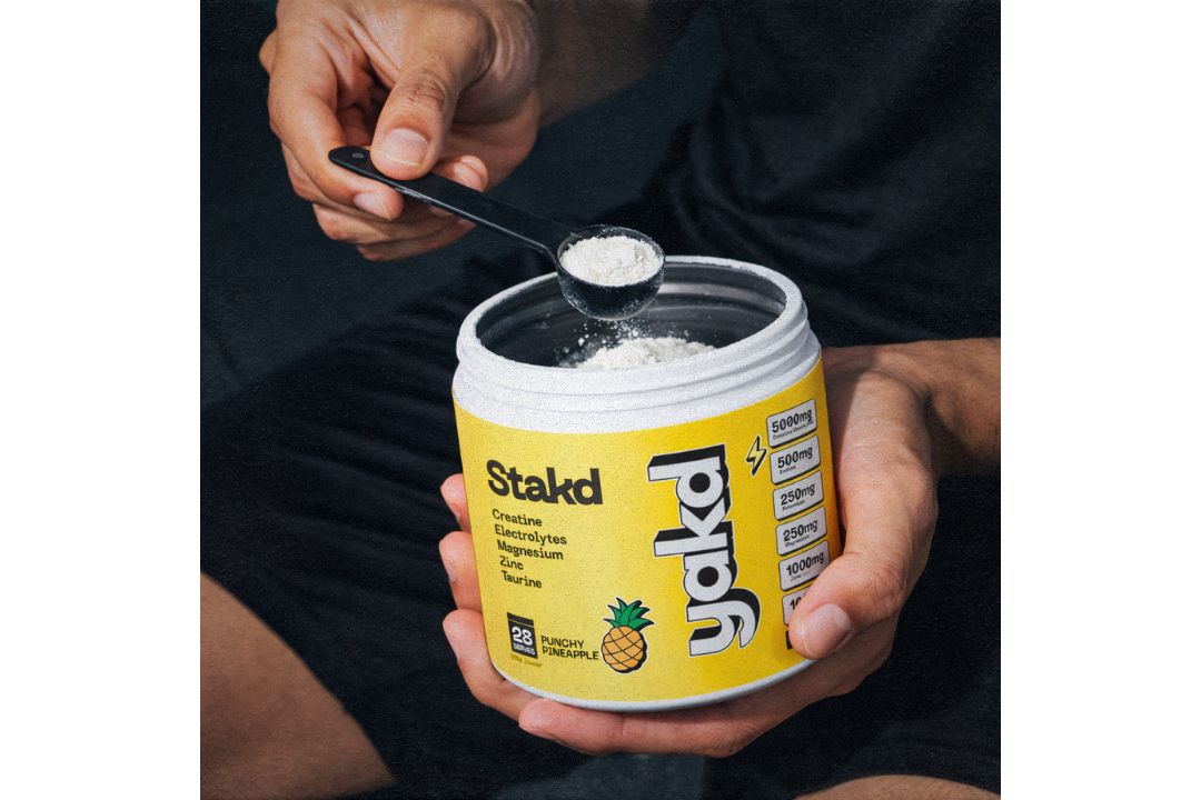 A person scoops a white powder supplement from a yellow container labeled 'Stakd'.
