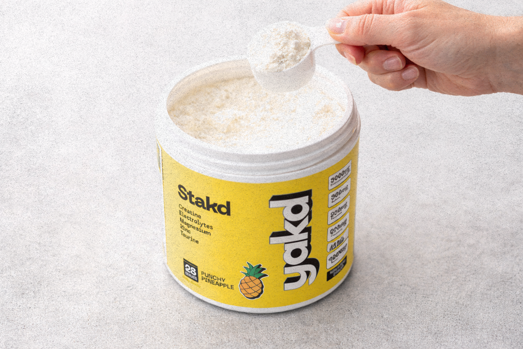 A hand holds a scoop of white powder above an open tub of Stakd supplement.