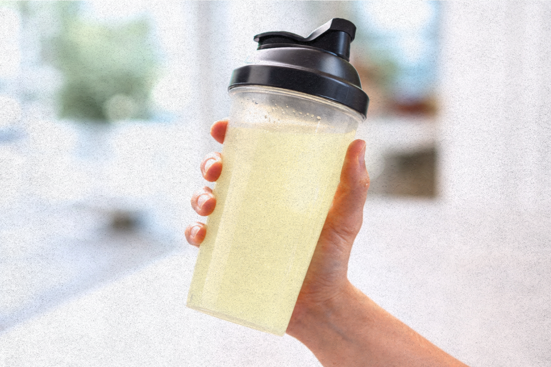 A hand holds a clear shaker bottle filled with a yellow beverage against a blurred background.