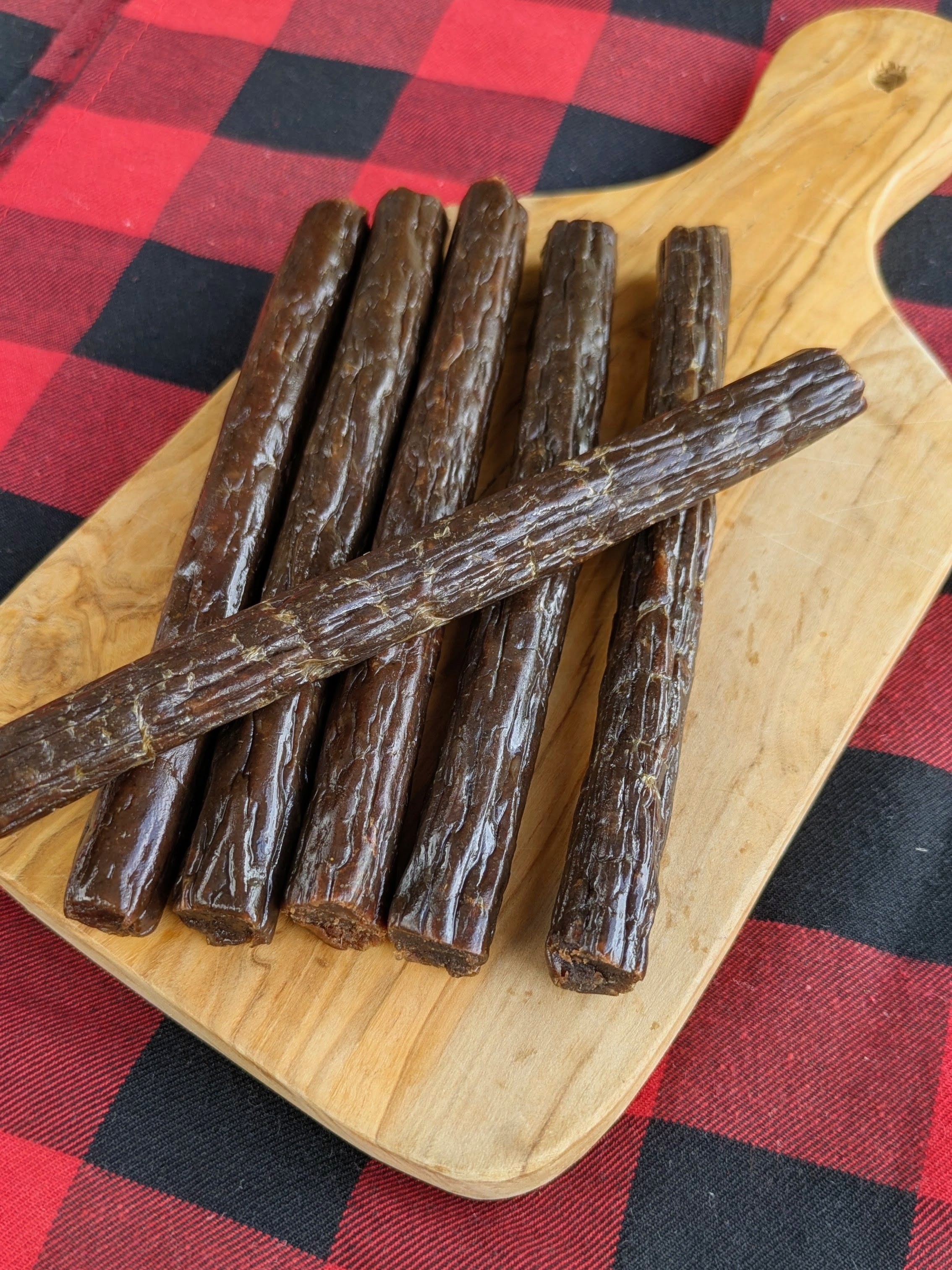 Wooden board with six beef sticks on a red and black checkered cloth.