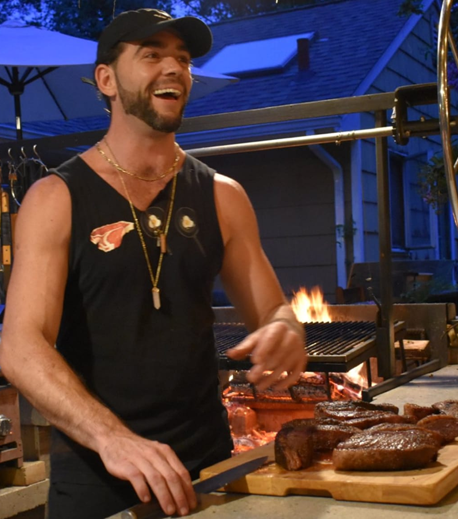 Man grilling steaks outdoors, smiling, with fire in the background.