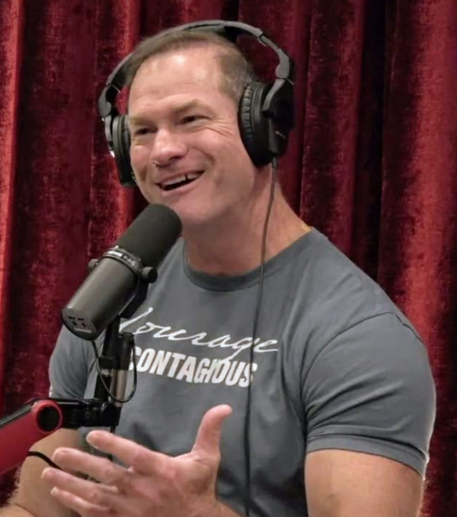 Man wearing headphones speaking into a microphone with a red curtain background.